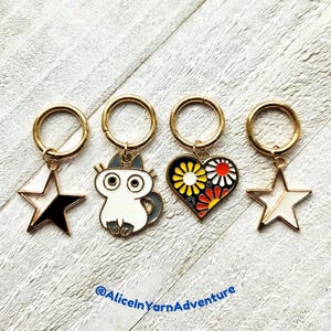 May include: Four gold stitch markers with different charms. The charms are a black and white star, a white cat with grey ears, a heart with a floral pattern, and a white star.