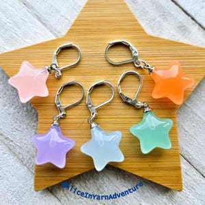Peut inclure: Cinq breloques en forme d'étoile dans différentes couleurs pastel, chacune attachée à un crochet d'oreille en argent. Les breloques sont en verre translucide et sont suspendues aux crochets.