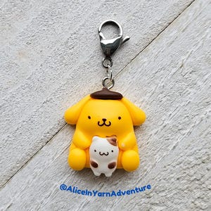 May include: A yellow and brown cartoon dog charm with a brown hat, holding a small white and brown cat charm. The charm is attached to a silver metal chain with a lobster clasp. The charm is on a white wooden background. @AliceInYarnAdventure
