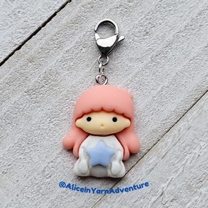 May include: A pink and white character charm with a blue star, attached to a silver metal lobster clasp. The character has pink hair and a white dress.