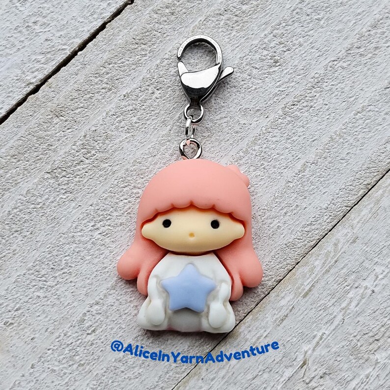 May include: A pink and white character charm with a blue star, attached to a silver metal lobster clasp. The character has pink hair and a white dress.