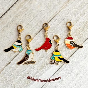 May include: Five bird-shaped charms with gold-coloured clasps. The birds are in various colours, including red, yellow, black, white, and brown. Some birds wear scarves or hats. The charms are on a light wood-grain surface.