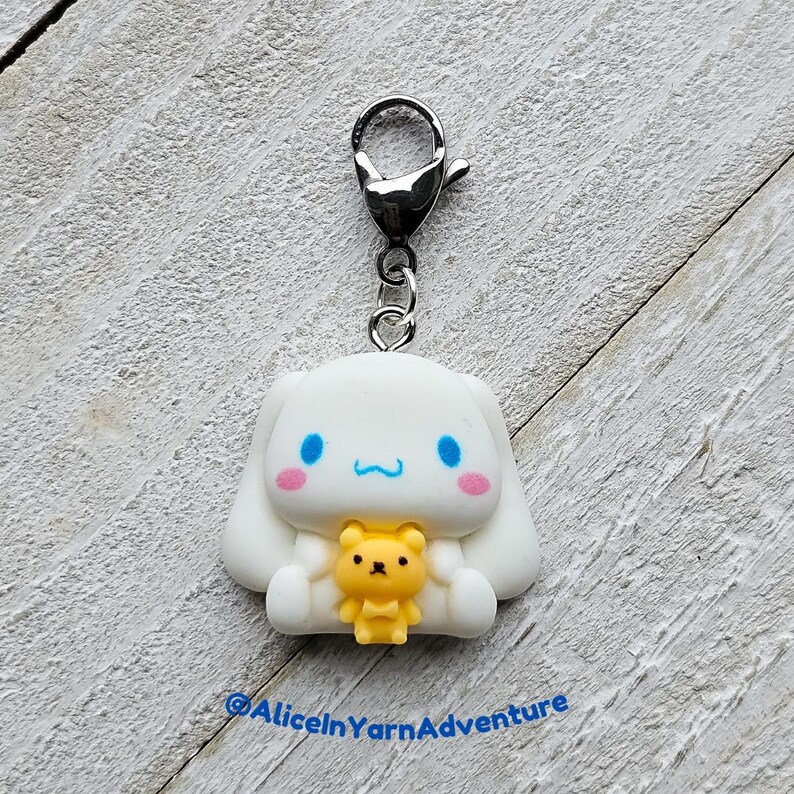 May include: White and blue character charm with a small yellow bear charm. The character has a blue bow and a pink nose. The charm is attached to a silver key chain.