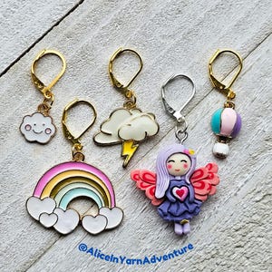 May include: Five colourful stitch markers with gold hoops. The markers feature a rainbow, a cloud with a smiley face, a cloud with lightning, a hot air balloon, and a fairy with pink wings.