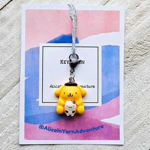 May include: A yellow and brown cartoon character keychain with a small white cat in its arms. The keychain is attached to a blue and white string. The keychain is on a pink and blue background with the text "Alice in Yarn Adventure" and "Keychain".