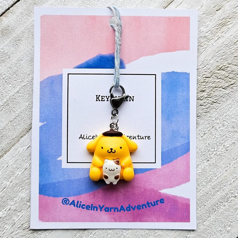 May include: A yellow and brown cartoon character keychain with a small white cat in its arms. The keychain is attached to a blue and white string. The keychain is on a pink and blue background with the text "Alice in Yarn Adventure" and "Keychain".