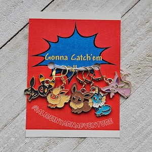 May include: Six colourful enamel charms featuring cartoon characters, attached to gold-coloured earring hooks. The charms are displayed on a red card with the text "Gonna Catch'em All" and "@ALICEINYARNADVENTURE".