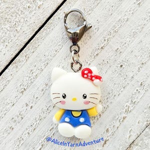 May include: A white Hello Kitty charm with a red bow and blue overalls. The charm is attached to a silver lobster clasp.