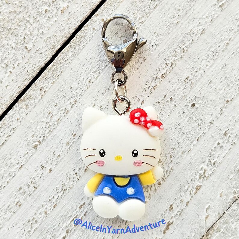 May include: A white Hello Kitty charm with a red bow and blue overalls. The charm is attached to a silver lobster clasp.