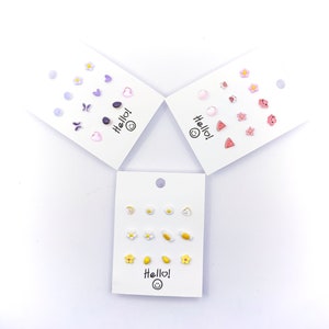 May include: Three sets of colourful stud earrings on white cards. Each set features a different theme: purple flowers, hearts, and butterflies; pink flowers, hearts, and watermelon slices; and yellow flowers, lemons, and eggs. Each card has the text "Hello!" with a smiley face.