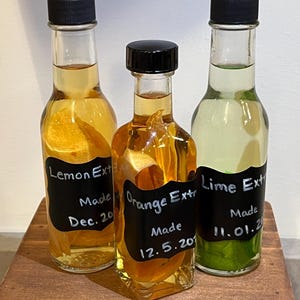 May include: Three glass bottles with black lids and handwritten labels. The bottles contain citrus fruit extracts. The labels read "Lemon Extract Made Dec. 20", "Orange Extract Made 12.5.20", and "Lime Extract Made 11.01.20".