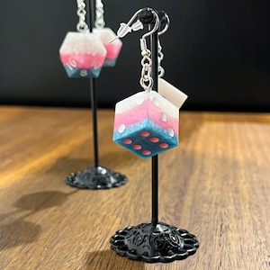May include: A pair of dice-shaped earrings. Each earring is a cube with white, pink, and blue layers, with red dots representing numbers. The earrings hang from silver-colored fish hook ear wires. The earrings are displayed on black stands.