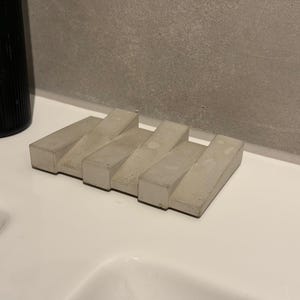 Può includere: Un portasapone in cemento grigio chiaro dal design moderno. Il portasapone presenta sei barre rettangolari disposte a gradini. Sullo sfondo è visibile una bottiglia verde scuro, appoggiata su una superficie bianca.
