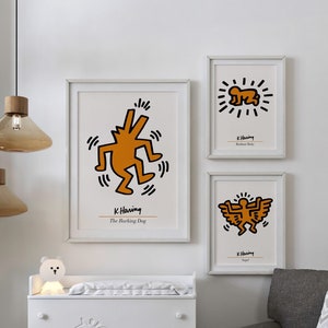 May include: A white dresser with four drawers features a giraffe design. The dresser is next to a white pouf with an orange center. Three framed prints with black outlines and orange figures are on the wall. The prints are by K. Haring and feature the titles "The Barking Dog", "Radiant Baby", and "Angel".
