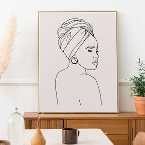 Peut inclure: Impression en ligne d'une femme avec un foulard, encadrée dans un cadre doré et accrochée à un mur blanc. L'impression est au-dessus d'une commode en bois avec un vase de pampas séchées et une bouteille en verre.