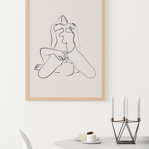 Peut inclure: Un dessin au trait noir et blanc d'une personne assise, les jambes crois&eacute;es. Le dessin est encadr&eacute; dans un cadre en bois clair et est accroch&eacute; &agrave; un mur blanc.