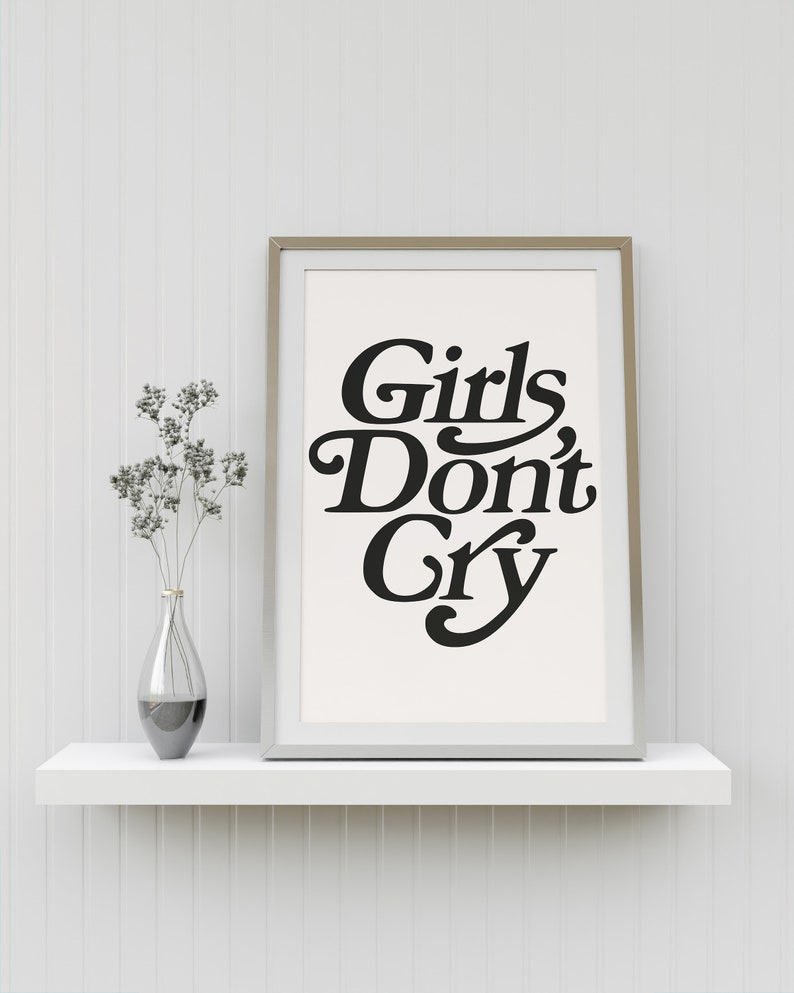 Puede incluir: Una impresi&oacute;n enmarcada con el texto "Girls Don't Cry" en una fuente en negrita y estilizada. La impresi&oacute;n est&aacute; sobre un fondo blanco y est&aacute; enmarcada en un marco plateado. La impresi&oacute;n est&aacute; sentada en un estante blanco contra una pared blanca.