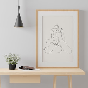 Peut inclure: Un dessin au trait noir et blanc d'une personne en position assise, encadr&eacute; dans un cadre en bois clair et accroch&eacute; &agrave; un mur blanc au-dessus d'un bureau en bois clair.