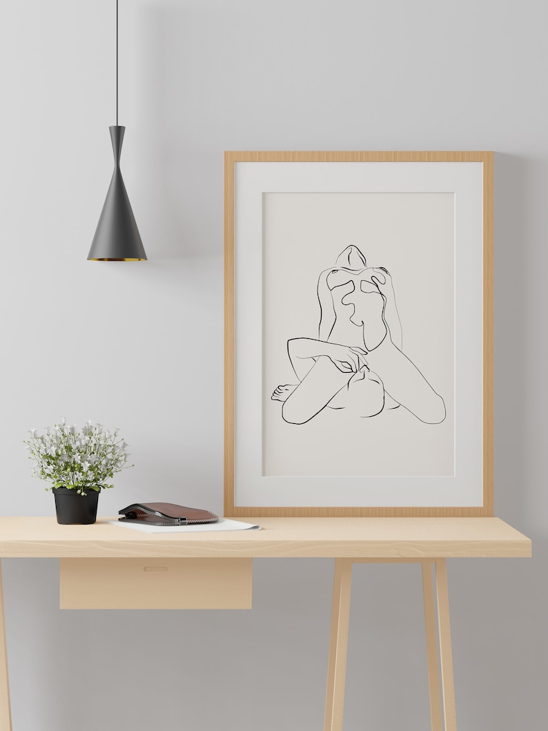 Peut inclure: Un dessin au trait noir et blanc d'une personne en position assise, encadr&eacute; dans un cadre en bois clair et accroch&eacute; &agrave; un mur blanc au-dessus d'un bureau en bois clair.