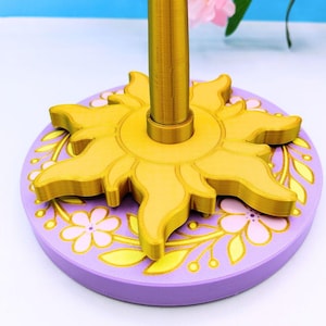 Puede incluir: Un soporte decorativo con un diseño de sol dorado sobre una base lavanda adornada con motivos florales. El soporte tiene un poste central dorado. El diseño evoca un tema de cuento de hadas.