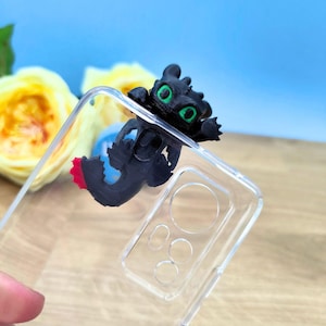 May include: A clear phone case with a black dragon figurine attached to the back. The dragon has green eyes and a red tail.