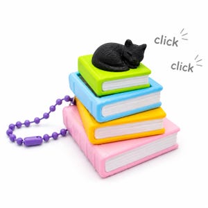 May include: A keychain featuring a stack of four miniature books in lime green, light blue, yellow, and pink, topped with a small black cat figurine. The books have white pages and the word click is printed twice near the cat with radiating lines.