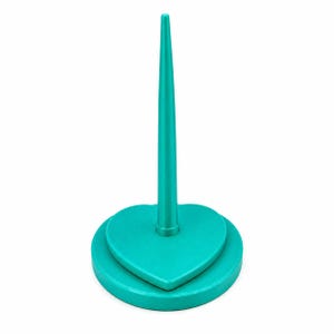 Op de afbeelding: Een helder talingkleurige ringhouder met een gladde, hartvormige basis en een lange, slanke, taps toelopende verticale staaf die vanuit het midden omhoog steekt. Het object heeft een egale kleur en een gladde, gepolijste afwerking.