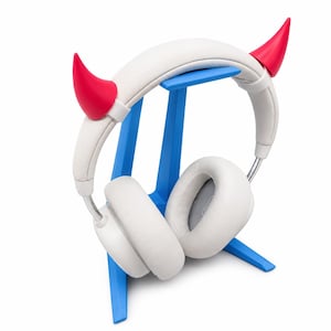 Könnte beinhalten: Weiße Over-Ear-Kopfhörer mit leuchtend roten Teufelshörnern am Kopfbügel, die auf einem blauen Kunststoffständer ruhen. Die Kopfhörer haben gepolsterte Ohrmuscheln und eine glatte weiße Oberfläche, während der Ständer ein schlichtes Design hat.