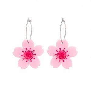 Puede incluir: Un par de pendientes de aro con colgantes de flores de color rosa claro de cinco pétalos. Cada flor tiene un centro de color rosa intenso con estambres radiantes. Los pendientes cuelgan de finos aros de metal plateado que contrastan.