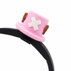 Puede incluir: Una diadema negra con un pequeño adorno de sombrero de copa rosa claro. El sombrero tiene un emblema de cruz blanca en la parte delantera y dos pequeños detalles rectangulares marrones a los lados, fijados sobre la banda lisa y oscura.