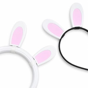 May include: Two headbands featuring upright rabbit ears. One headband is white with white and pink ears, while the other is a thin black band with white and pink ears. The ears are rounded and feature a soft pink center surrounded by a white border.