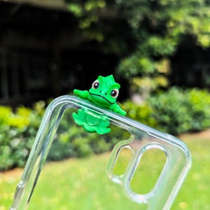 Può includere: Una custodia trasparente per telefono con una decorazione a forma di rana verde brillante. La rana ha grandi occhi ed è posizionata sull'angolo della custodia. Lo sfondo è sfocato, mostrando un fogliame verde.