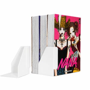 Op de afbeelding: Een set van drie mangaboeken getiteld Nana deel 1 door Ai Yazawa, met een felroze omslag met twee geïllustreerde personages, rechtop gehouden door twee witte plastic boekensteunen. De boeken staan naast elkaar met zichtbare paginaranden.