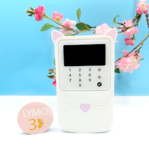 Puede incluir: Un dispositivo de punto de venta (TPV) blanco con un diseño de cara de gato, con un teclado y una pantalla negra. Un objeto circular rosa con el texto "LYMO 3" está al lado del dispositivo. Flores rosas y un fondo azul completan la imagen.