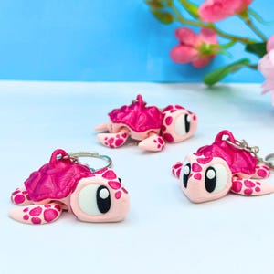 May include: Three handmade turtle keyrings. Each keyring features a pink and white turtle design with a textured shell and a silver keyring. The turtles have large, expressive eyes and are set against a blue and white background.