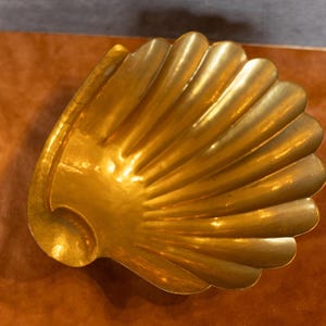 May include: A gold-colored metal seashell-shaped dish. The dish has a smooth, shiny surface and is designed to resemble a real seashell.