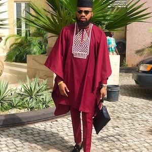 May include: A man wearing a maroon traditional African outfit with a geometric pattern on the chest. He is wearing a black cap, sunglasses, and black loafers. He is holding a black clutch bag.