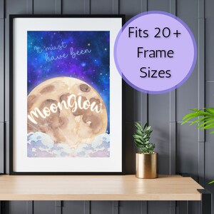 Könnte beinhalten: Ein Aquarelldruck eines Vollmonds mit dem Text "It must have been Moonglow" in einem schwarzen Rahmen. Der Druck befindet sich auf einem Holztisch mit einer Pflanze in einem goldenen Topf.