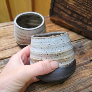 May include: Two ceramic cups with a rounded shape and a ribbed texture. The upper portion of each cup features a speckled off-white glaze, while the lower portion is finished in a solid matte black glaze, creating a distinct two-tone appearance.
