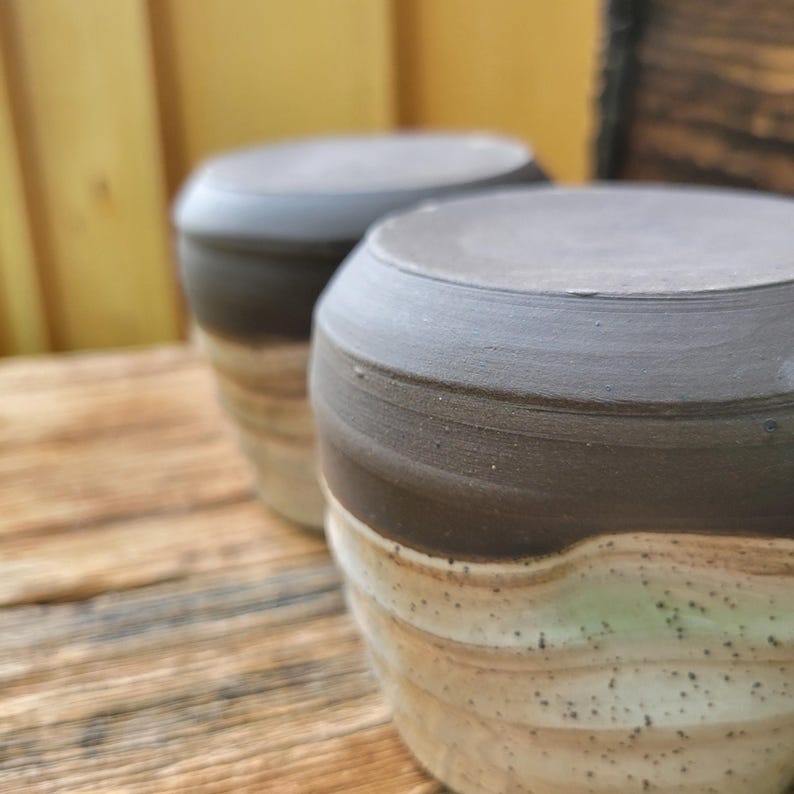 May include: Two ceramic cups featuring a dark charcoal gray rim and a textured, speckled cream-colored base with subtle horizontal ridges. The cups have a cylindrical shape with a smooth, matte finish on the upper portion and a slightly uneven surface.
