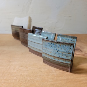May include: A row of four ceramic sponge holders with a curved, open-front design. Each holder features a distinct glaze, ranging from dark brown to light blue and speckled teal, with a natural clay base visible at the bottom of each piece.