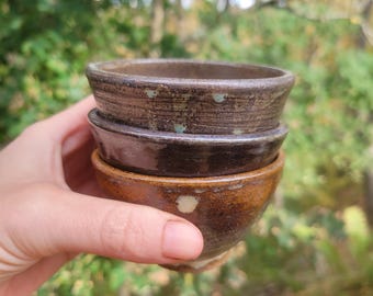 May include: A stack of three small ceramic bowls with a glazed finish. The top bowl is a textured grey-brown, the middle bowl is a smooth dark brown, and the bottom bowl is a warm ochre-brown with a lighter cream-colored spot near the base.