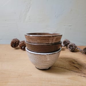 May include: A stack of three ceramic bowls with a speckled finish. The top bowl is light brown, the middle bowl is a darker brown, and the bottom bowl is white with dark speckles. Each bowl features a rounded shape and a textured, rustic surface.
