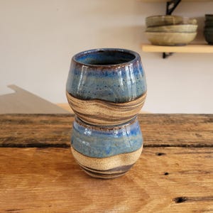 May include: Two stacked ceramic cups featuring a rounded, bulbous shape. The upper portion of each cup is glazed in a glossy, mottled blue, while the lower section displays a natural, earthy beige and brown marbled clay texture with a smooth finish.