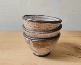 May include: A stack of three small ceramic bowls with a speckled tan and white glaze. Each bowl features a dark brown rim and a rounded base, showcasing a hand-thrown appearance with visible horizontal lines and subtle color variations in the clay.