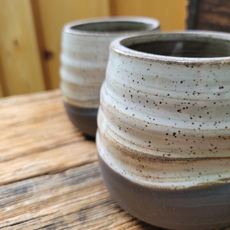 May include: Two ceramic tumblers featuring a rounded, ribbed silhouette. The upper portion of each vessel is finished in a speckled off-white glaze, while the lower section is a solid, dark brown matte finish, creating a distinct two-tone appearance.