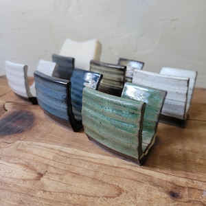May include: A collection of U-shaped ceramic sponge holders featuring horizontal ribbed textures. The holders are finished in various glossy glazes, including shades of green, blue, black, and white, with some showing speckled or dark brown edges.