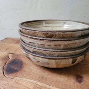 May include: A stack of three round ceramic bowls featuring a speckled cream and beige glaze with a dark brown rim. The bowls have a textured, hand-thrown appearance with visible variations in the glaze and a wide, shallow basin design.