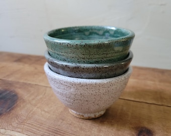 May include: A stack of three small ceramic bowls with a speckled finish. The top bowl is a vibrant green, the middle bowl is a dark charcoal gray, and the bottom bowl is a light cream color. Each bowl features a rounded shape and a smooth rim.