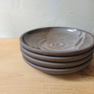 May include: A stack of four handmade ceramic plates. The plates are a dark brown color with a lighter brown swirl pattern in the center. The plates have a slightly rough texture and a simple, rustic design.